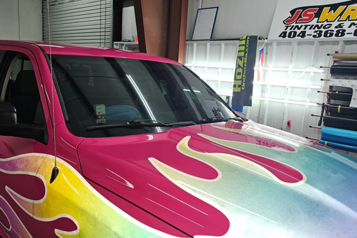 A pink truck with flames painted on the hood is parked in a garage.