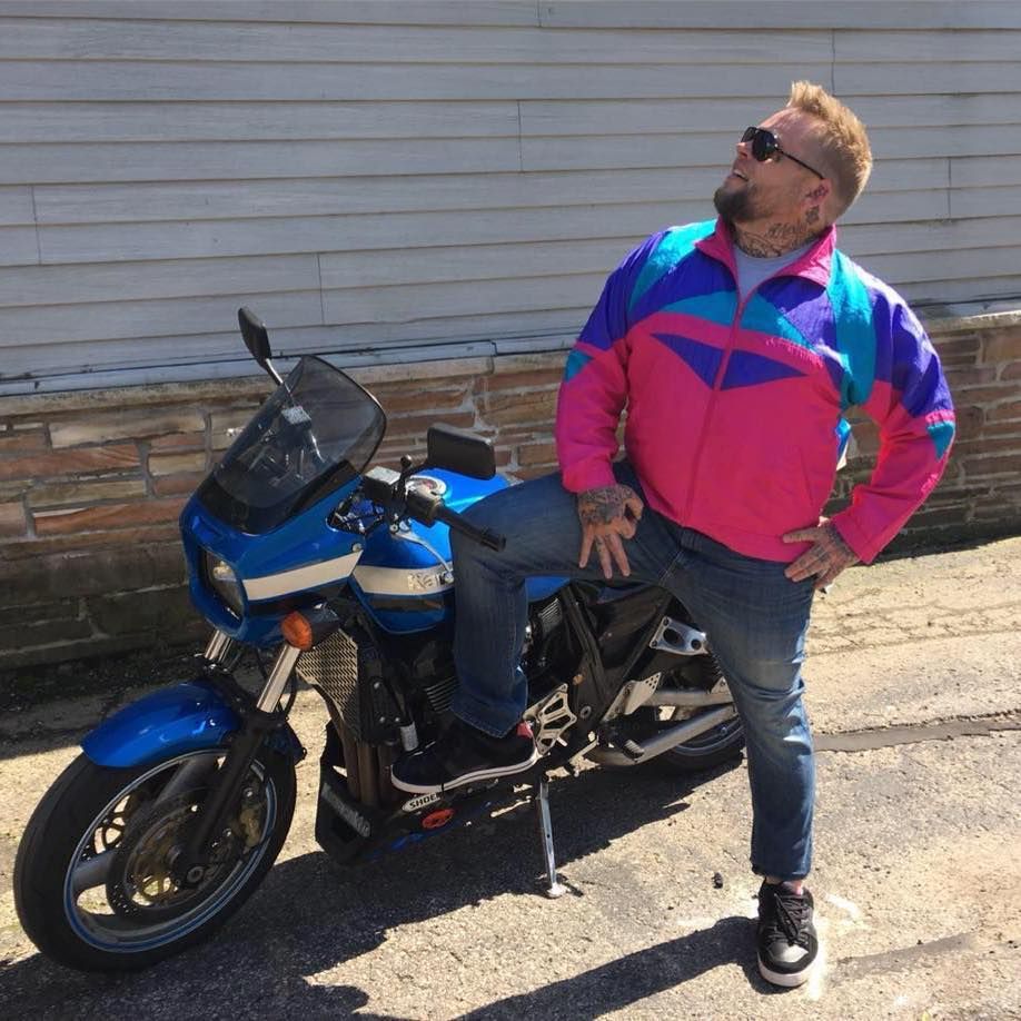 A man in a pink and blue jacket leans on a blue motorcycle