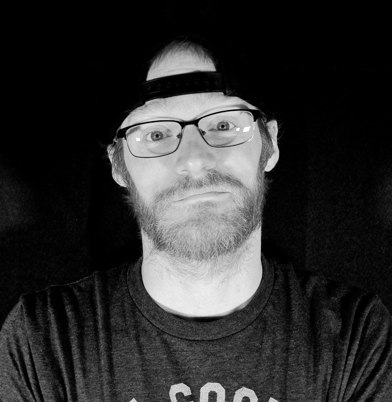 A man with a beard and glasses is wearing a hat and a t-shirt.
