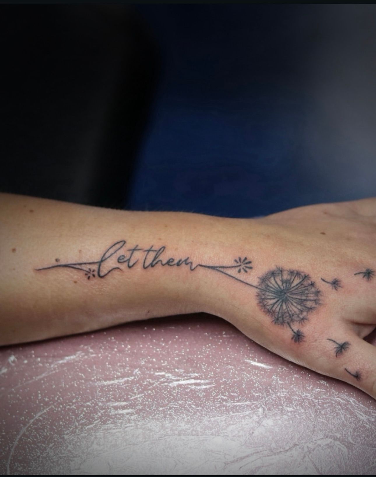 A woman has a tattoo of a dandelion and the words let them on her wrist.