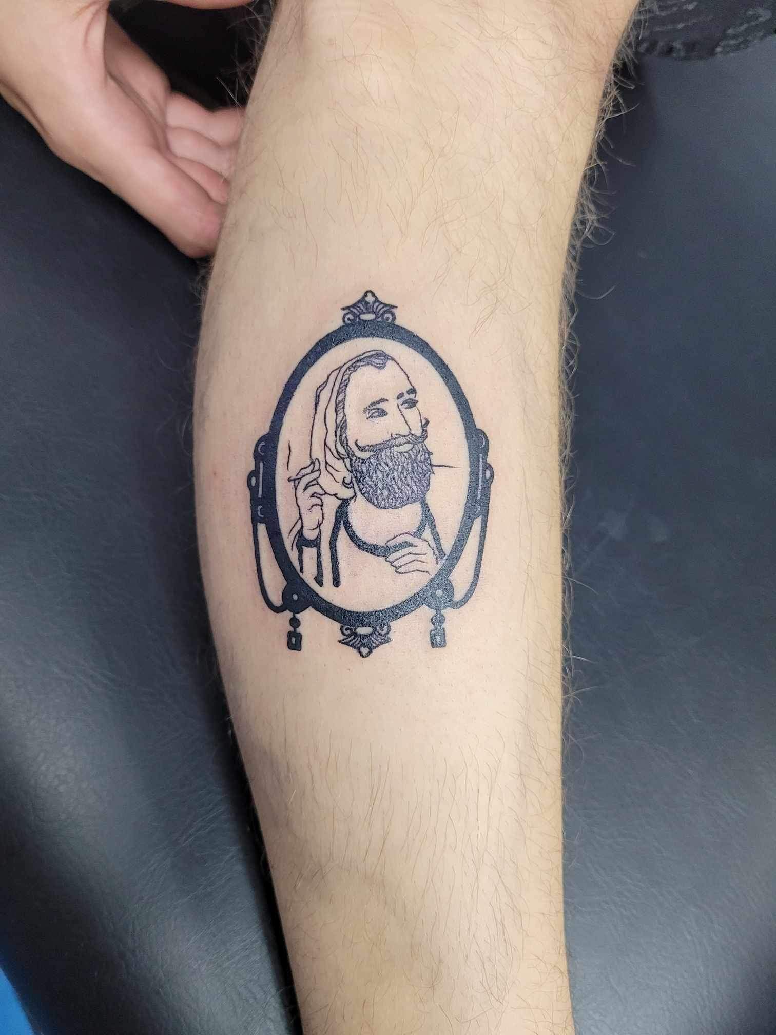 A person has a tattoo of a man with a beard in a mirror on their leg.