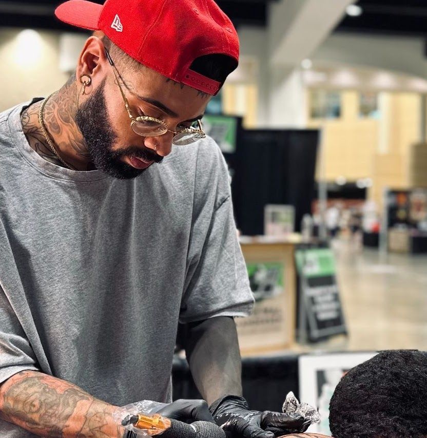 A man in a red hat is getting a tattoo on another man 's arm.