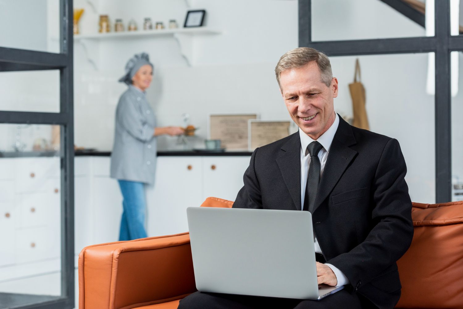 A person in a suit sits on a couch using a laptop, while another person works in a kitchen in the background.