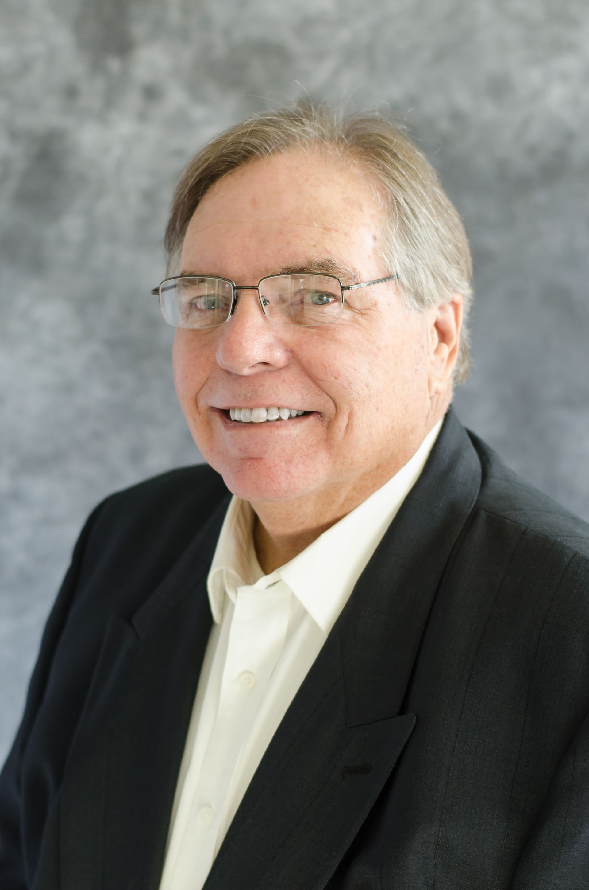 A person wearing glasses and a dark blazer over a light shirt, smiling in front of a gray mottled backdrop.