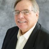 A person with glasses, wearing a white shirt and a dark blazer, smiles against a gray, textured background.