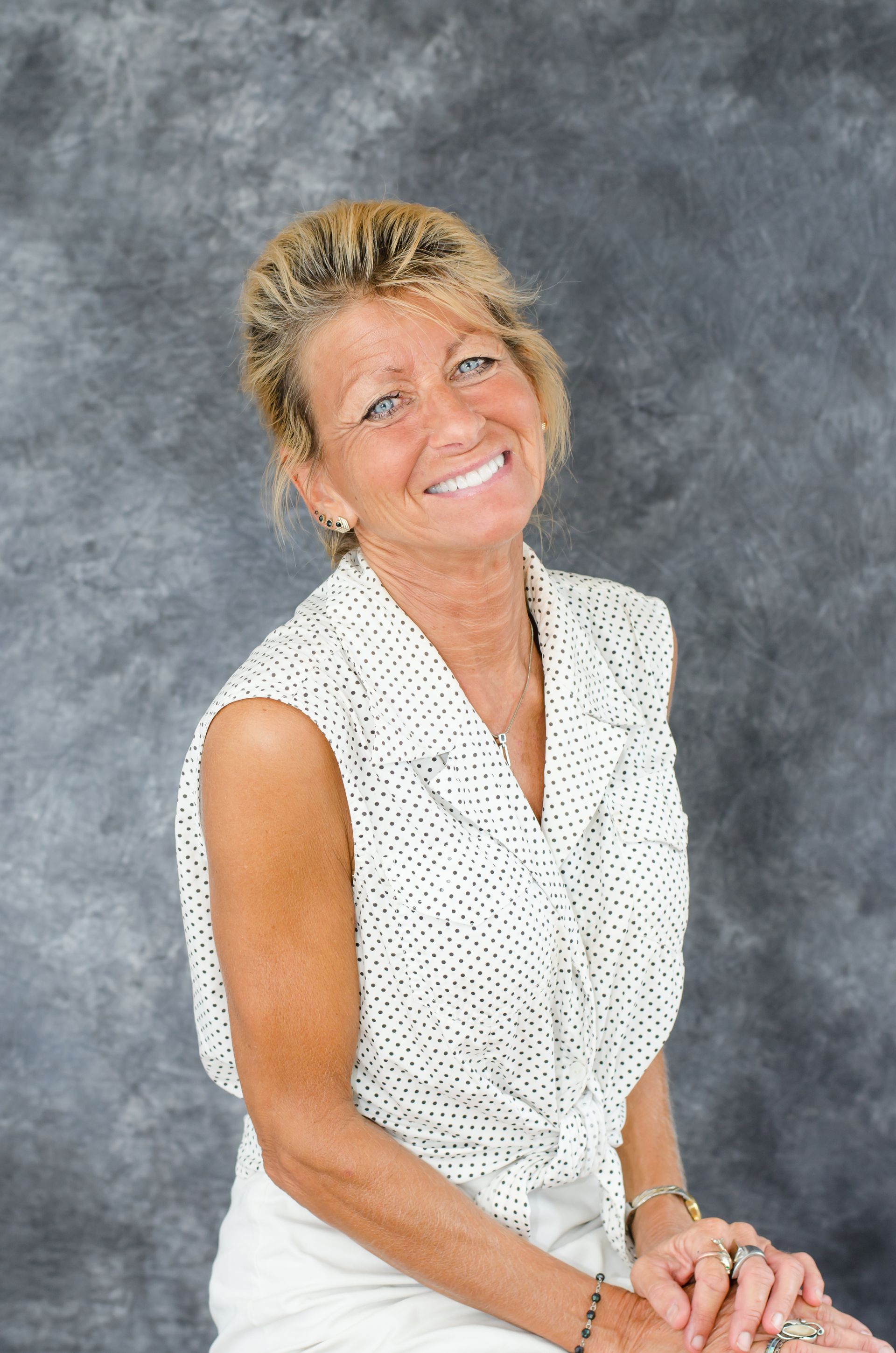A person with blonde hair in a white polka-dot blouse smiles while sitting against a mottled gray backdrop.