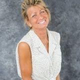 A smiling woman with blonde hair, wearing a white sleeveless top with black polka dots against a gray mottled background.