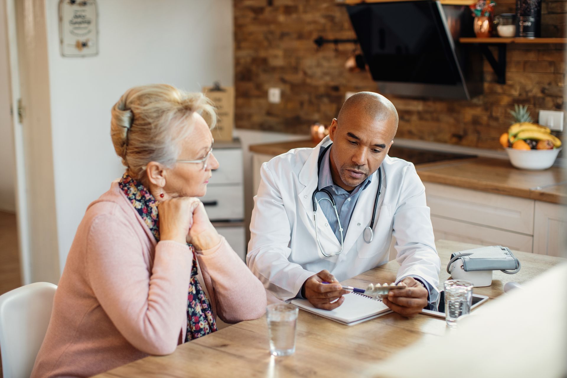 A medical professional in a white coat shows a blister pack of pills to a patient during a home health consultation.