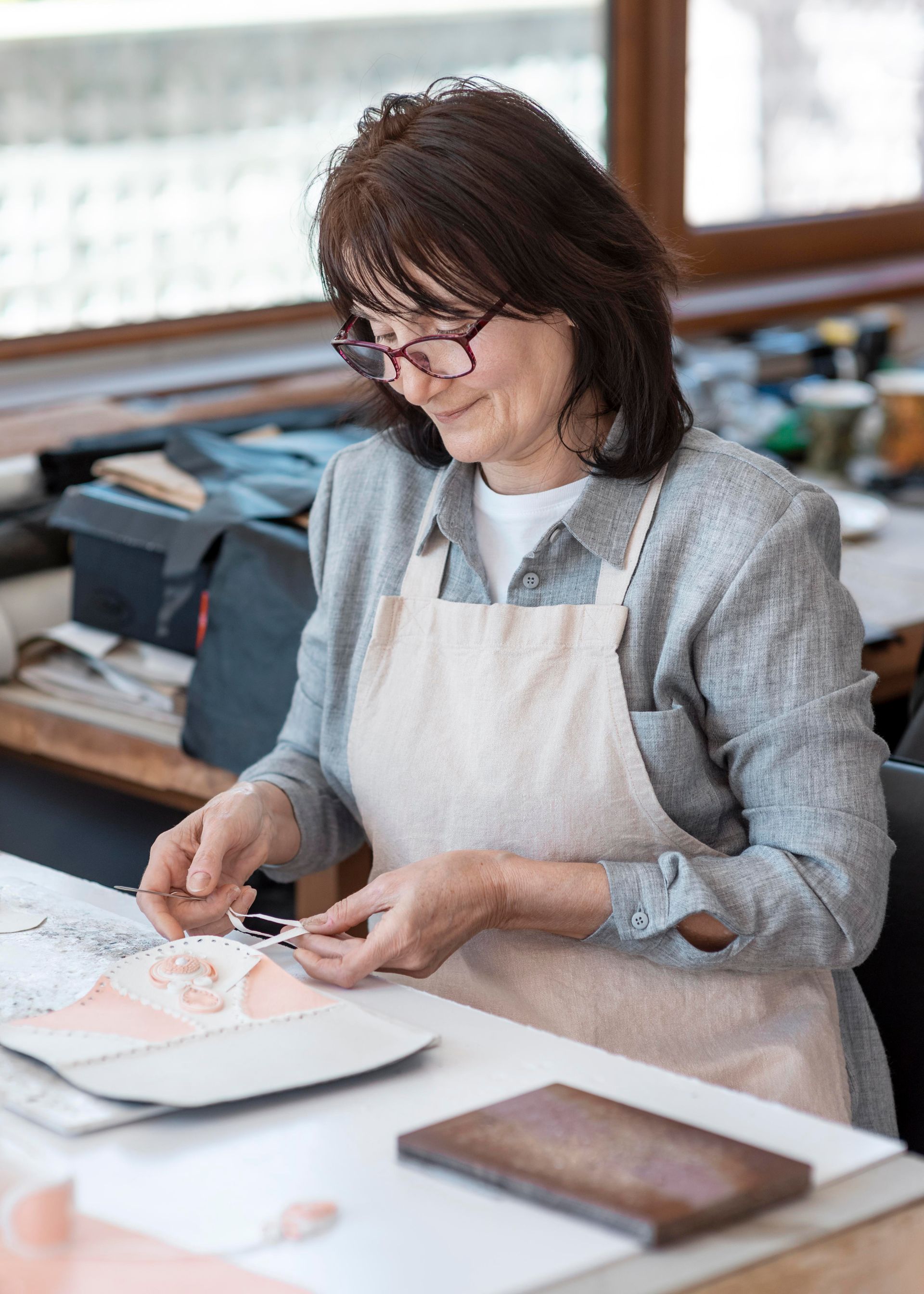 A person wearing glasses and an apron works on a detailed craft project at a desk in a well-lit studio.