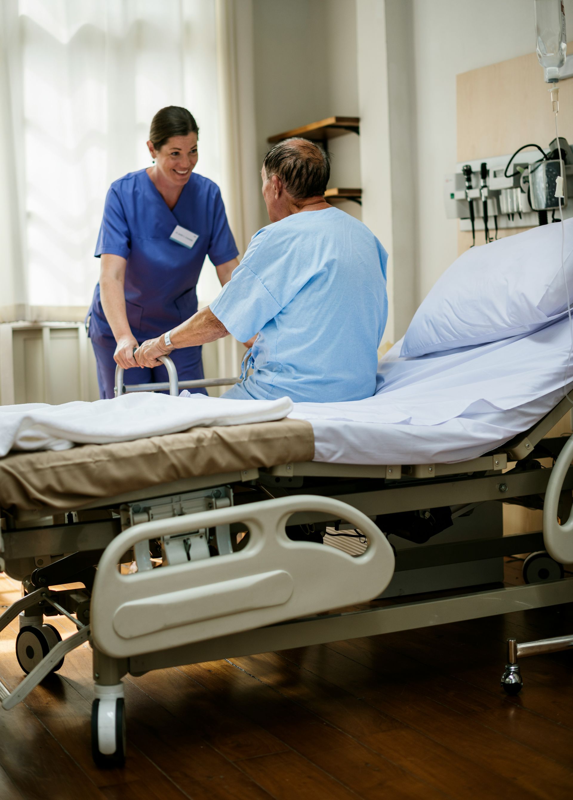 A healthcare worker in blue scrubs helps a patient sit up on the side of a hospital bed in a bright, modern room.