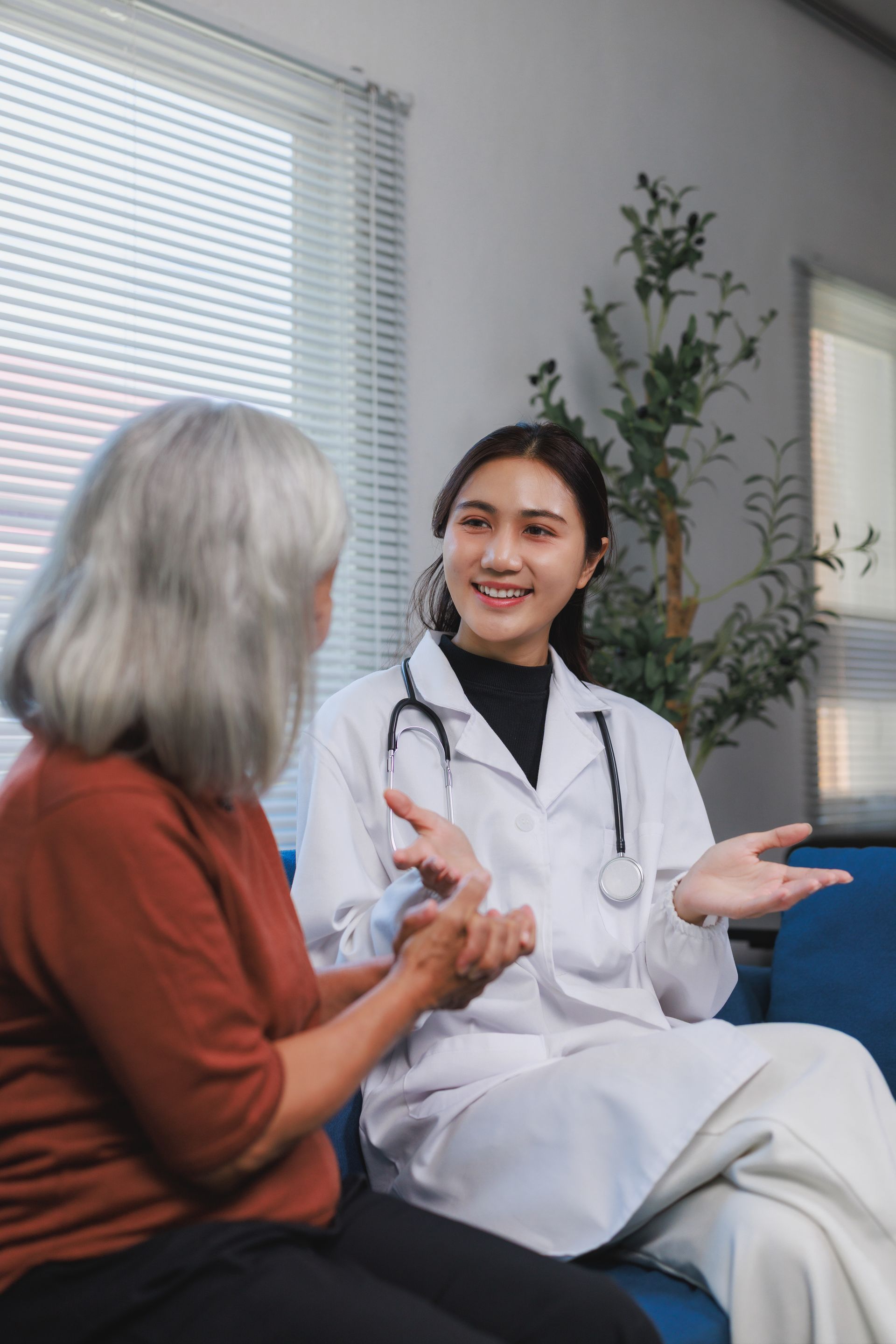 A medical professional in a white coat with a stethoscope talks and gestures to a patient in a bright, indoor setting.