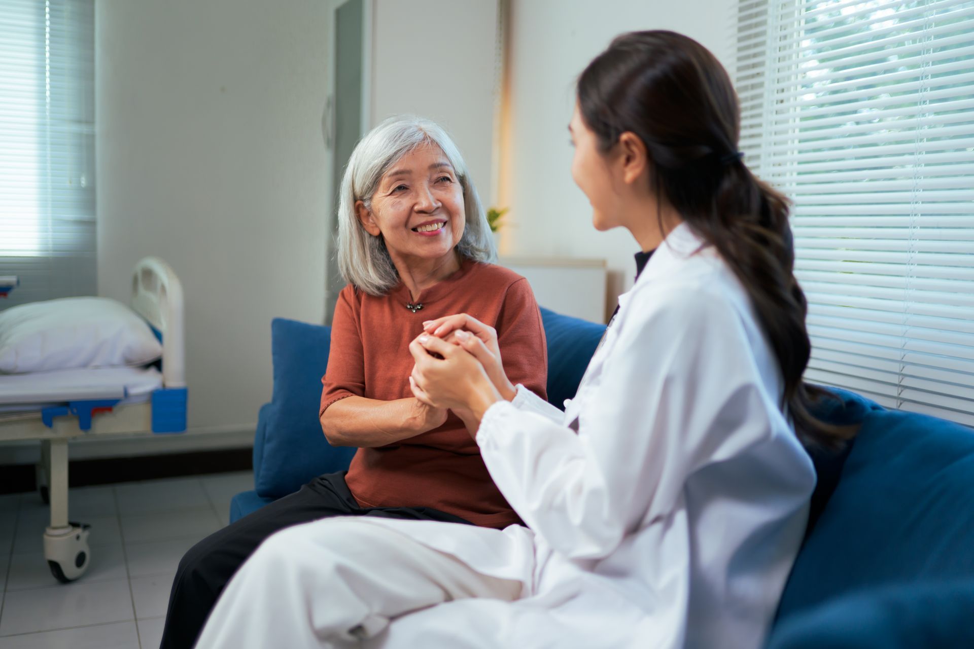 A medical professional wearing a white coat holds an elderly patient's hands, providing comfort in a clinical room.