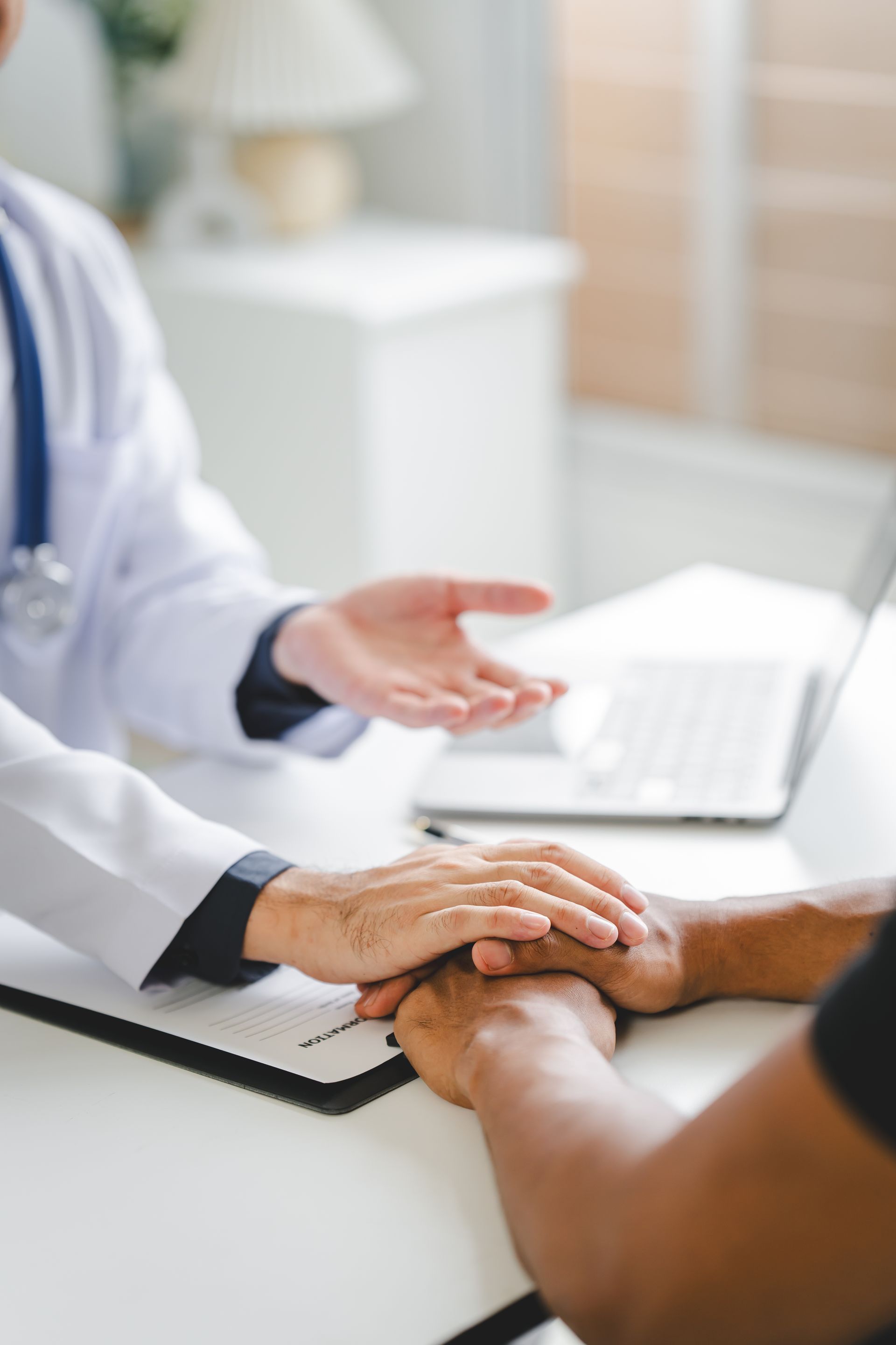 A doctor wearing a white coat and stethoscope holds a patient's hand in a clinical office to offer support and comfort.