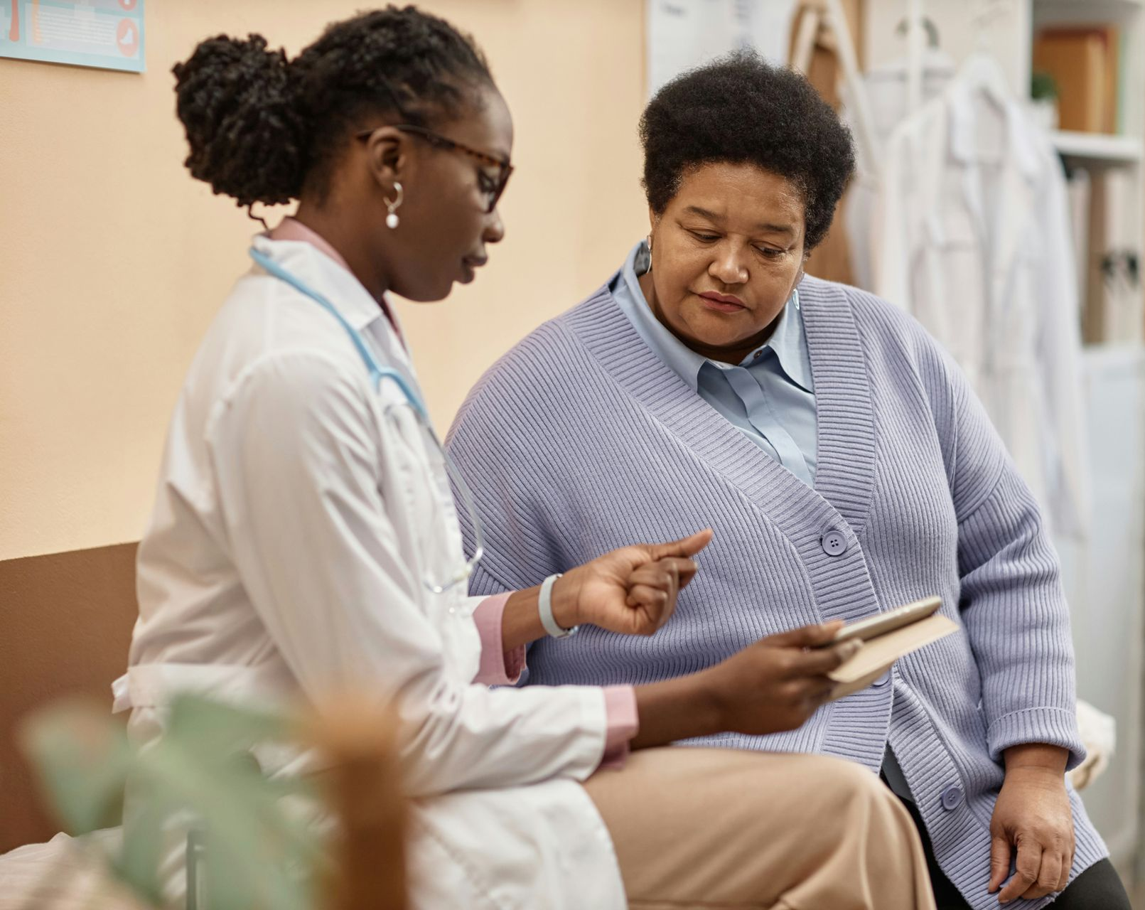 A healthcare provider in a lab coat and stethoscope shows a tablet to a patient in a medical office.