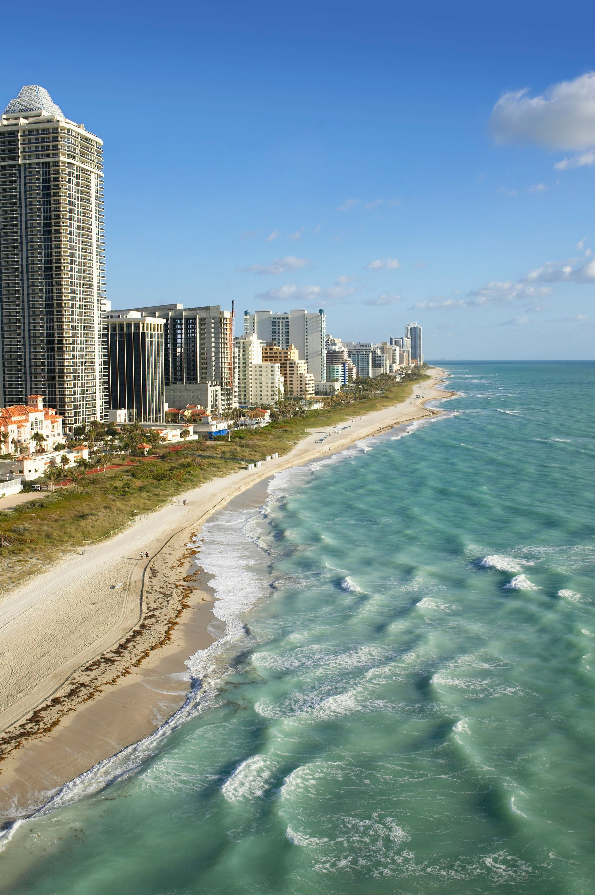 A high-angle view of a sandy beach coastline bordering tall apartment buildings against a clear blue sky.