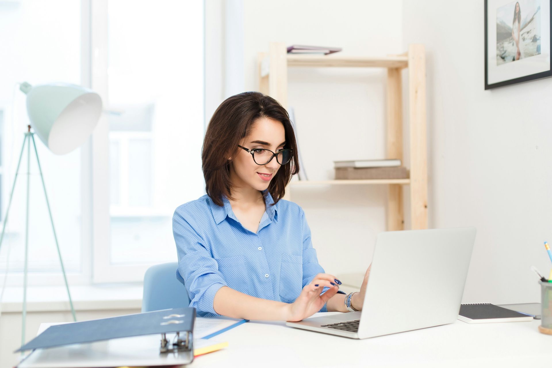 A person in a blue button-down shirt and glasses working on a laptop at a bright, minimalist desk.