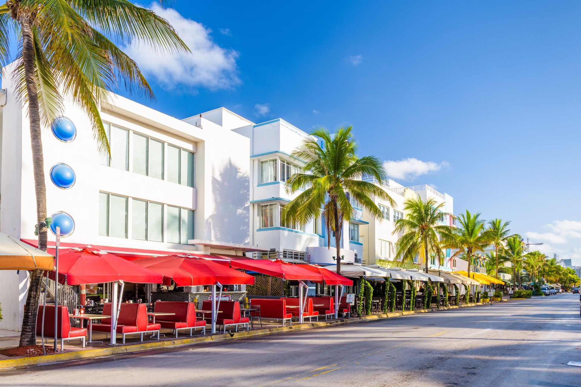 Art Deco buildings on Ocean Drive, Miami, with palm trees and red outdoor restaurant seating under a clear blue sky.