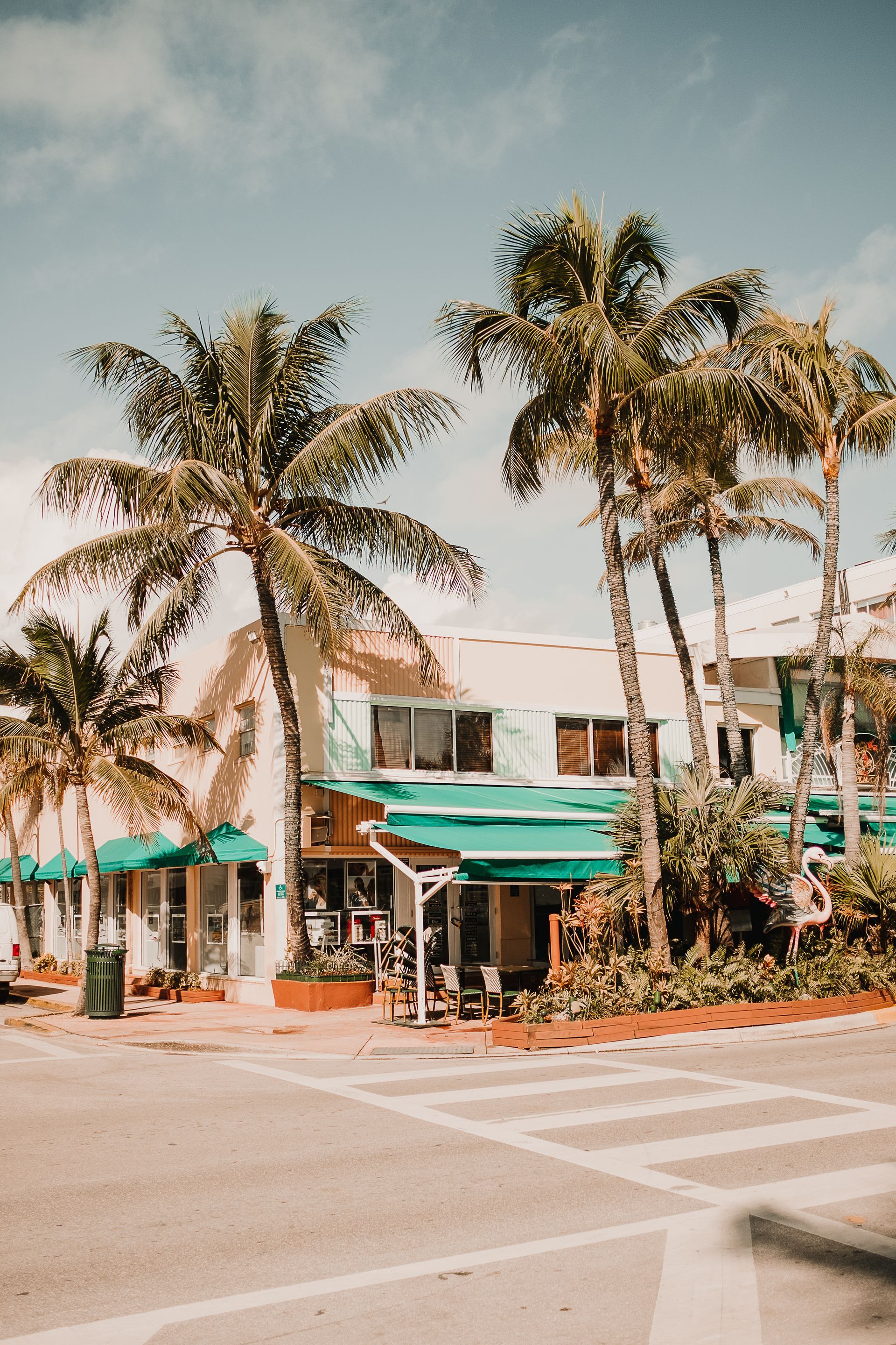 A cream-colored building with green awnings sits on a sunny street, surrounded by tall palm trees.
