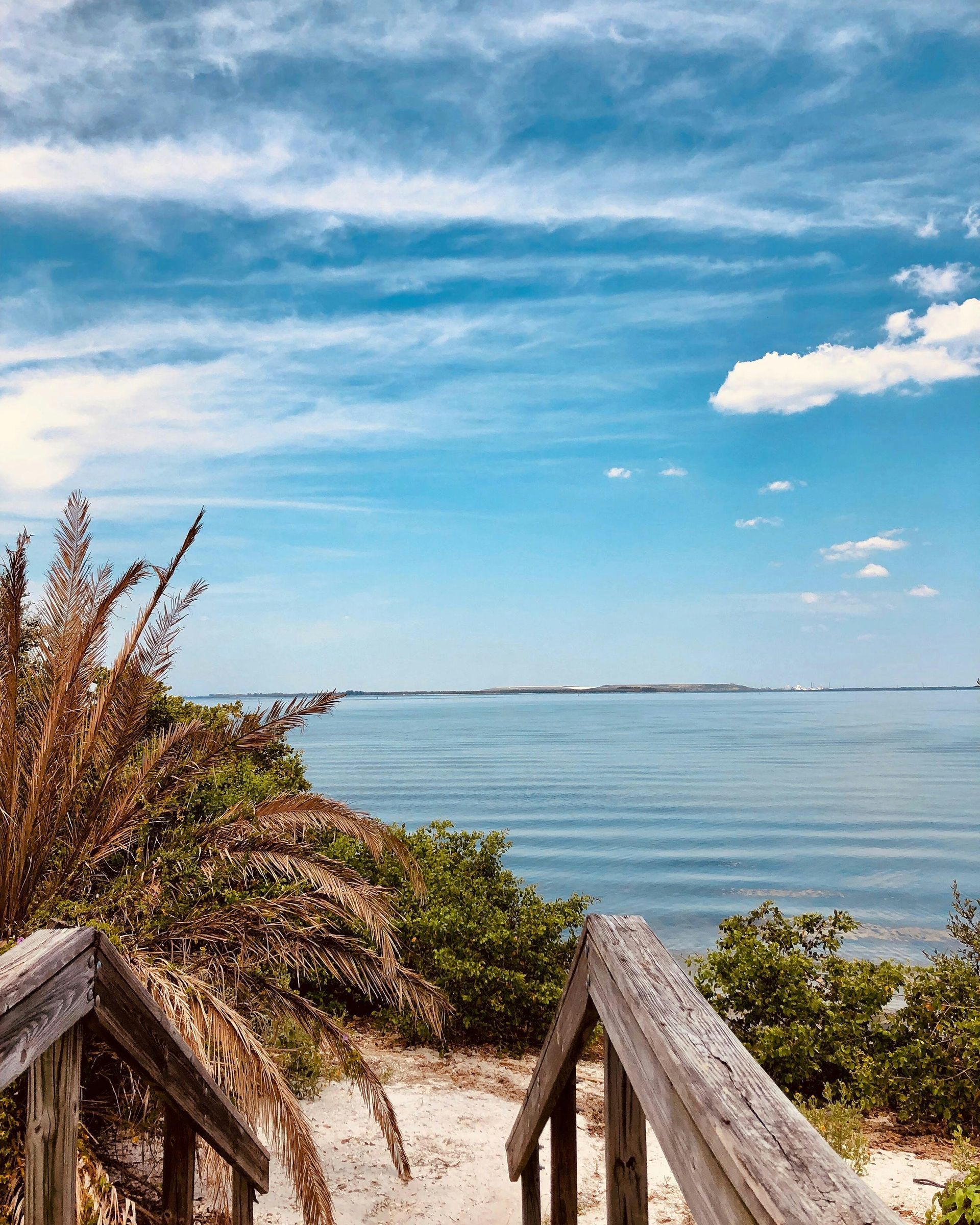 Wooden stairs lead down a sandy path toward a calm, blue bay under a bright, partly cloudy sky.