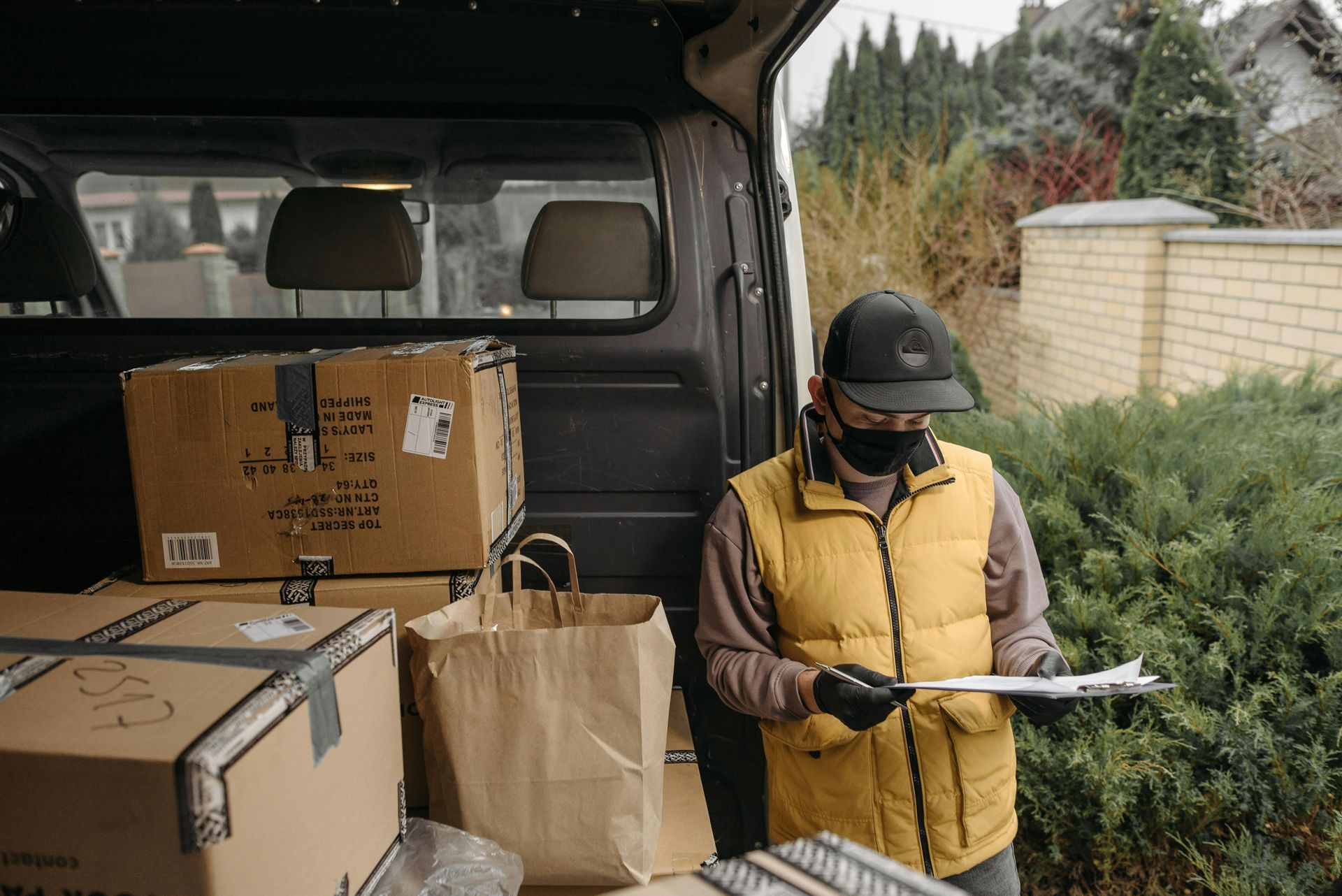 A masked delivery person in a yellow vest checks a clipboard while standing by a van filled with boxes and bags.