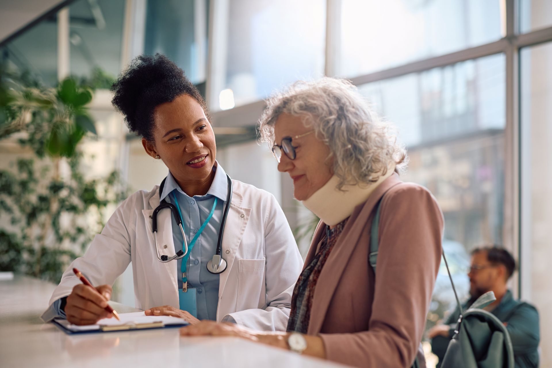 A medical professional in a white coat talks to a patient wearing a neck brace while writing on a clipboard at a desk.