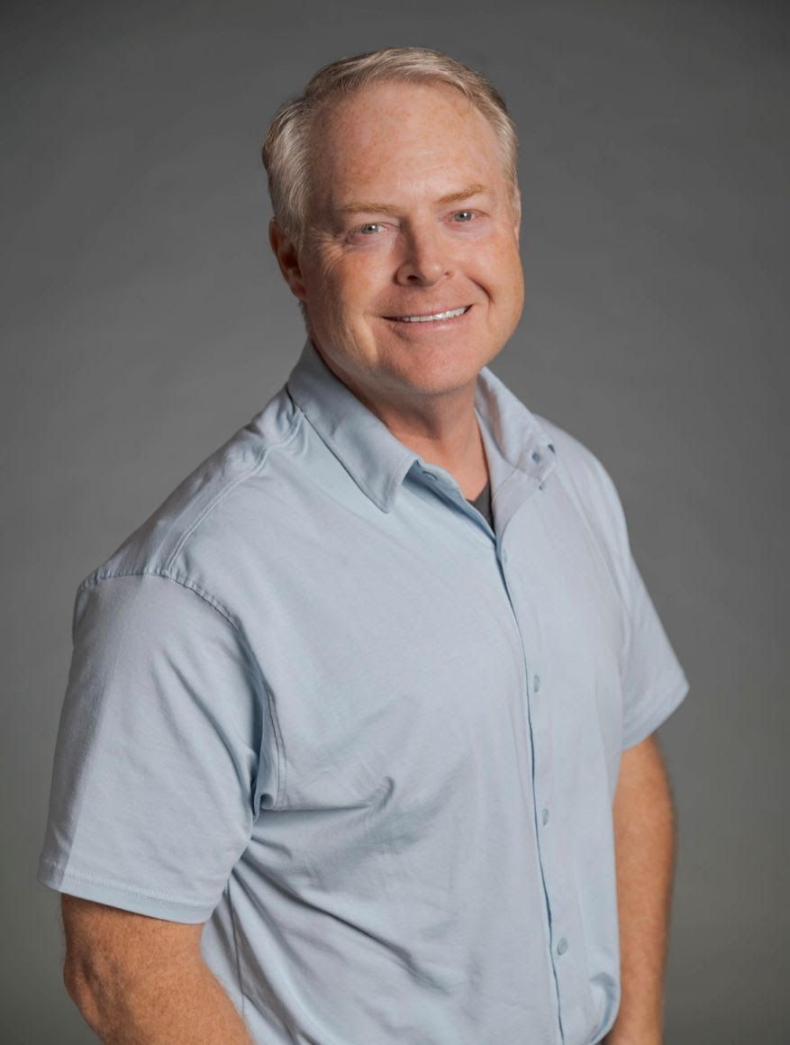 A person wearing a light blue short-sleeved button-up shirt smiling against a neutral gray background.