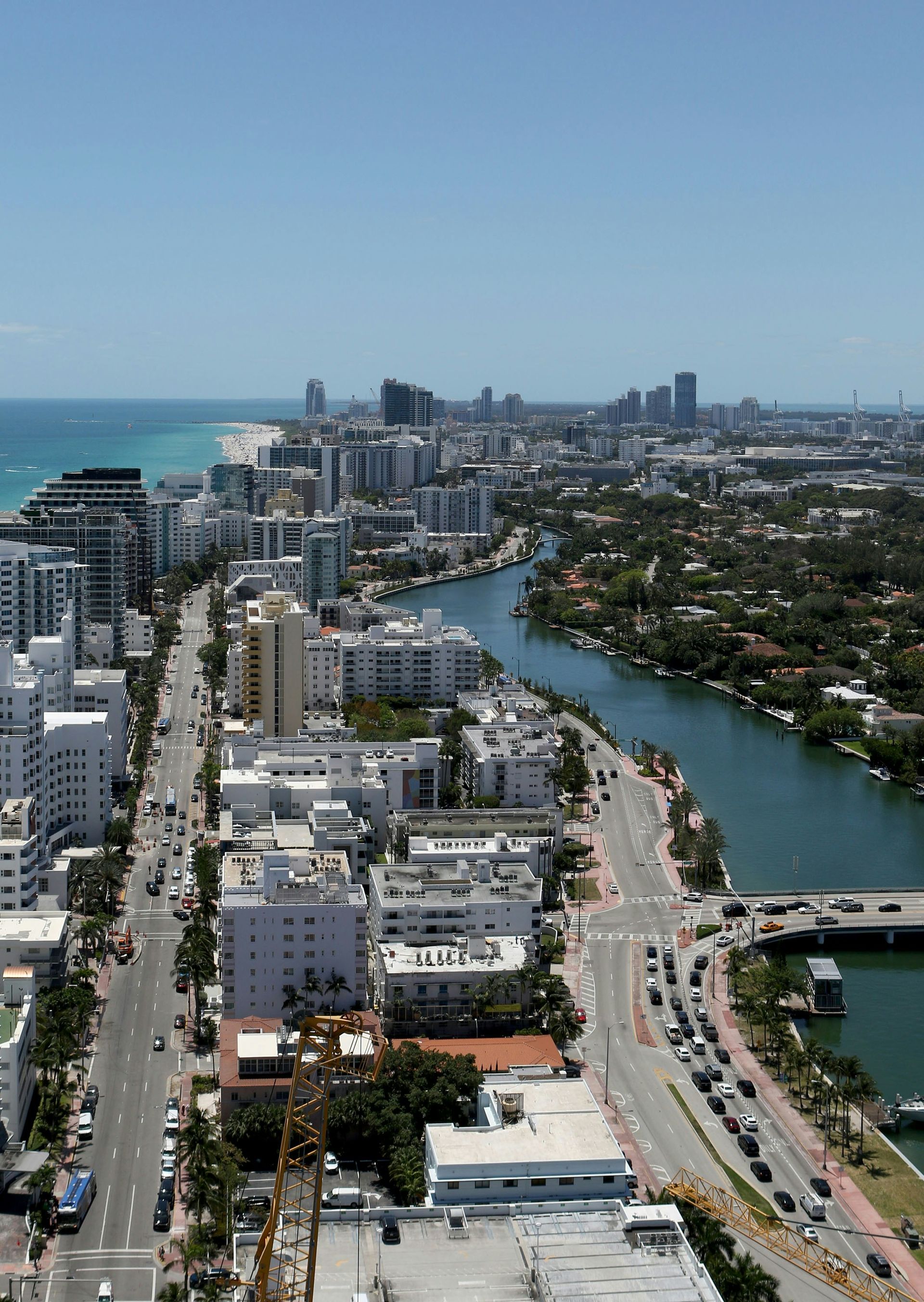 An aerial view of a Miami beach district featuring buildings, a canal, palm trees, and the ocean in the distance.