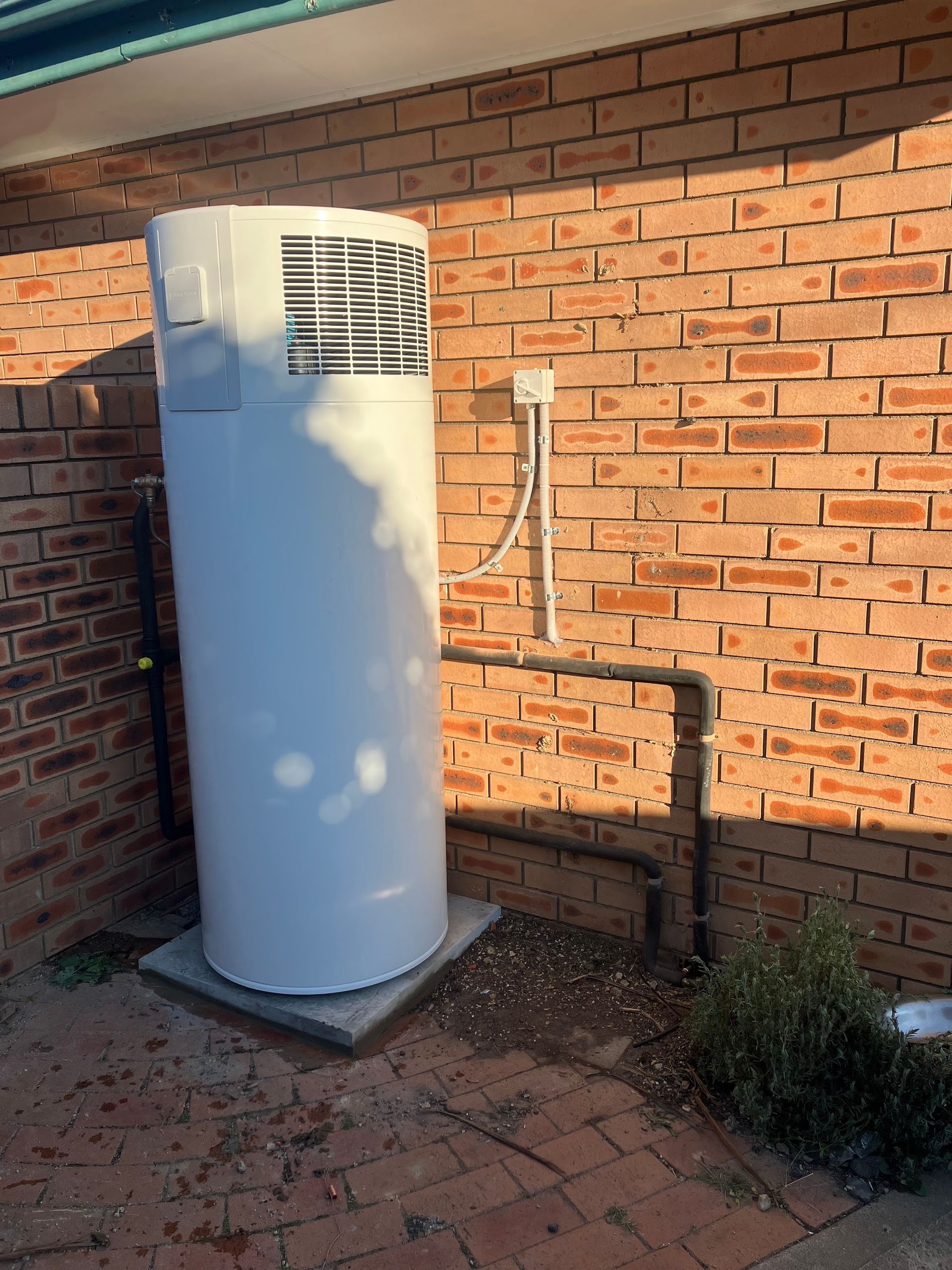 White outdoor unit beside a brick wall, with utility piping in a sunlit patio area — Plumbing in Canberra, ACT