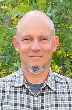 A bald man with a beard is wearing a plaid shirt and smiling for the camera.