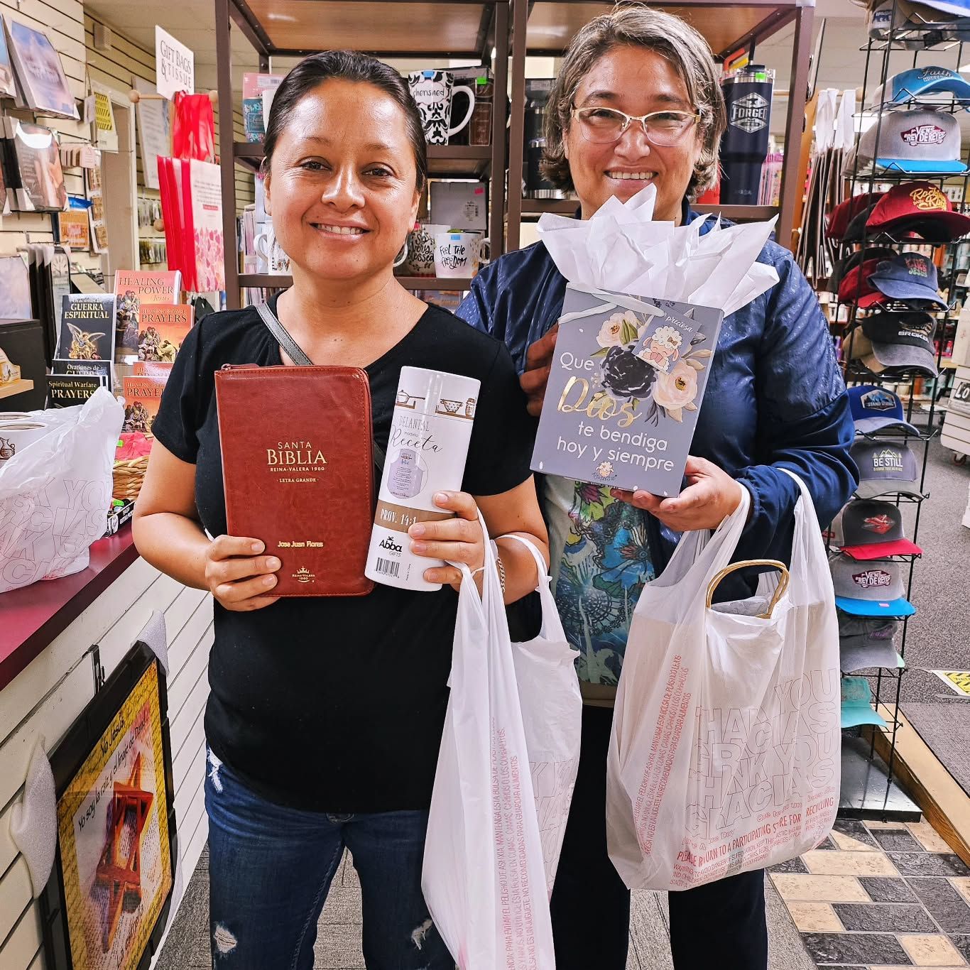 Two women smiling, holding gifts and shopping bags inside a gift shop.