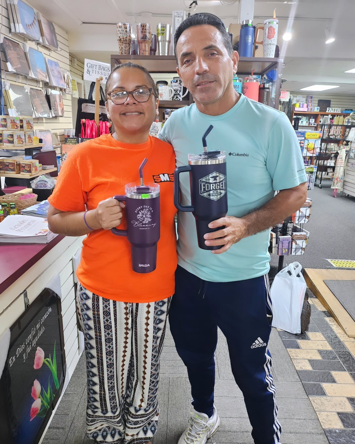Two people in a store holding large mugs. Woman in orange shirt, man in blue, smiles. Products on shelves in background.