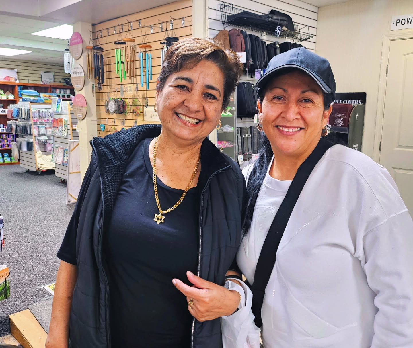 Two smiling women pose in a store. One points to her chest, the other wears a hat.