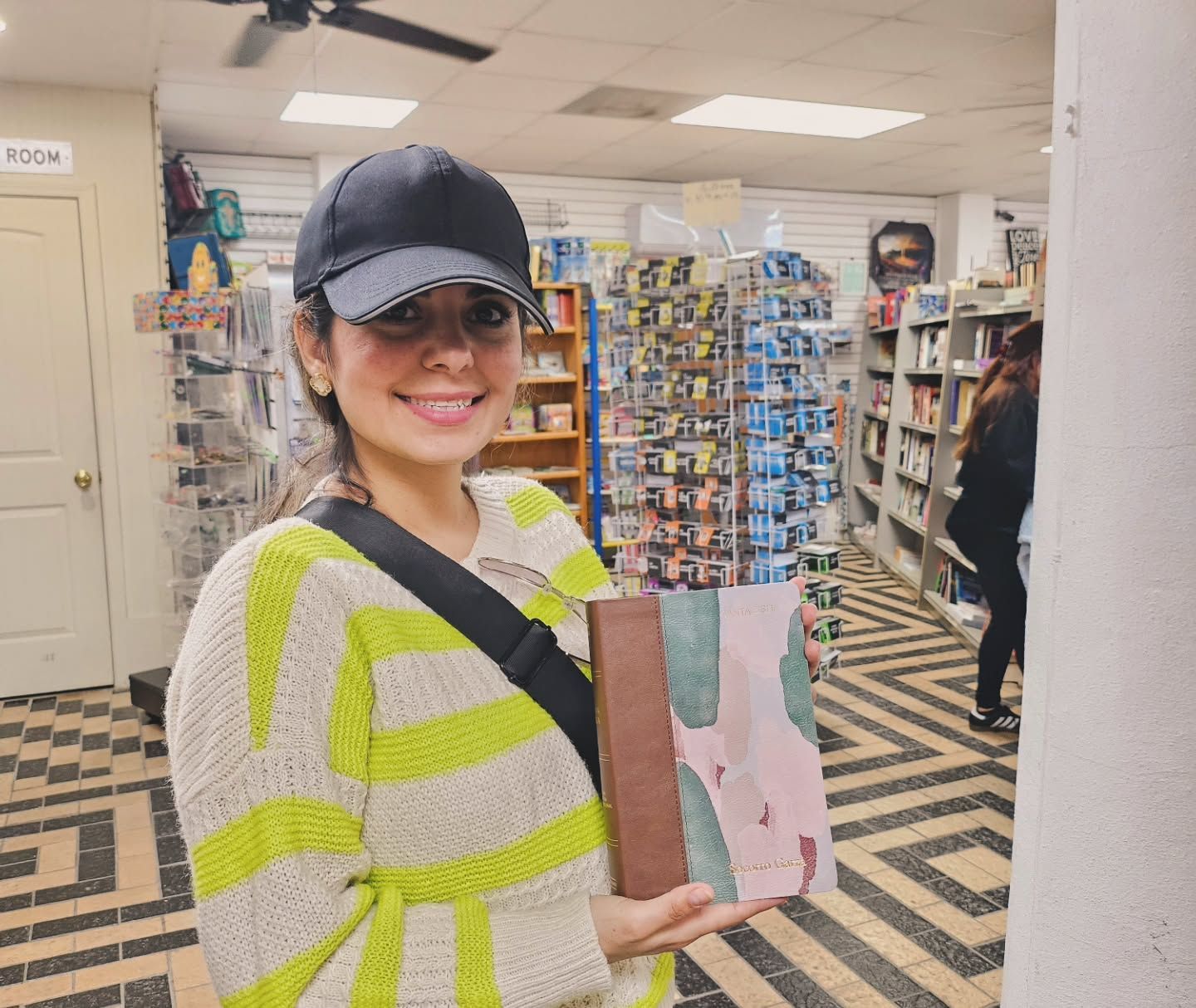 Woman in bookstore holding a book, smiling. Wearing hat and sweater. Bookshelves in background.