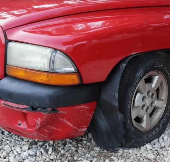 A red truck with a damaged bumper is parked on gravel