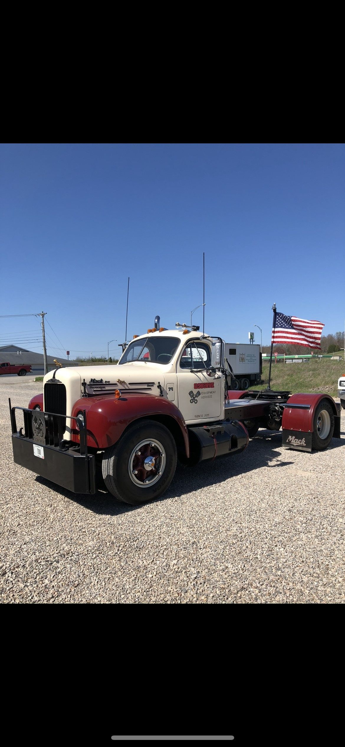 An antique white and maroon semi-truck parked on gravel under a blue sky, an American flag waving in the background.