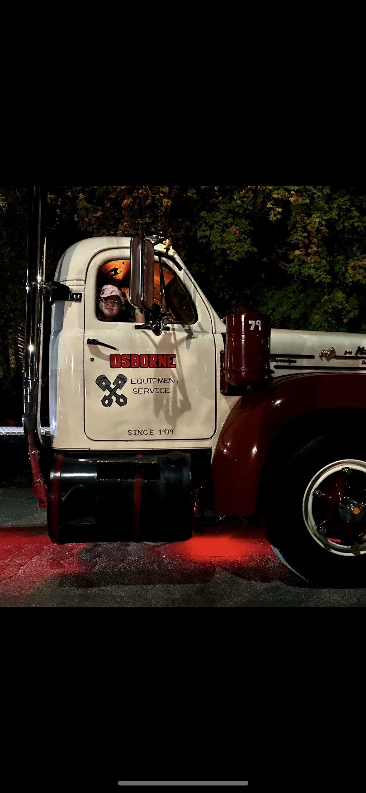 A vintage white and burgundy semi-truck with red underglow lighting. A driver is visible inside the cab.
