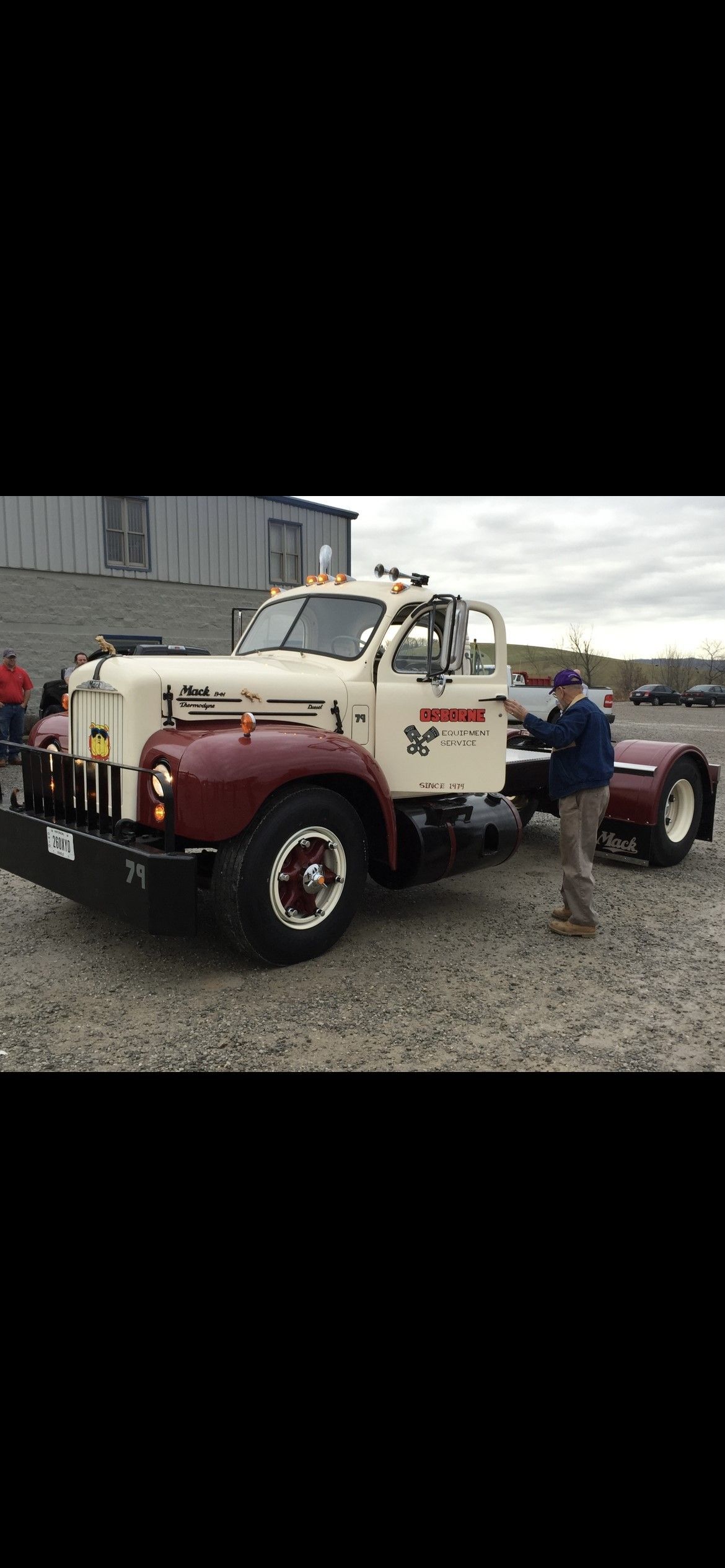 A vintage white and burgundy Mack truck parked on gravel with a person standing next to it.