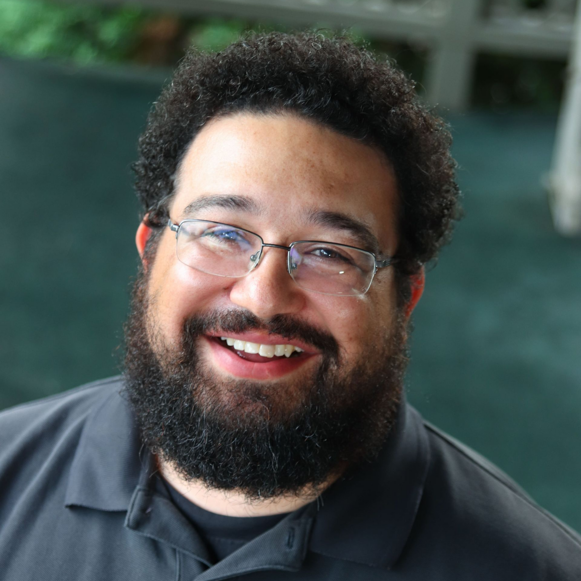 A smiling person with a beard and glasses wearing a dark collared shirt