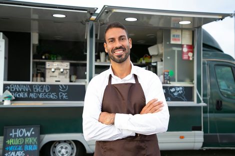 Man in apron stands smiling in front of a food truck.