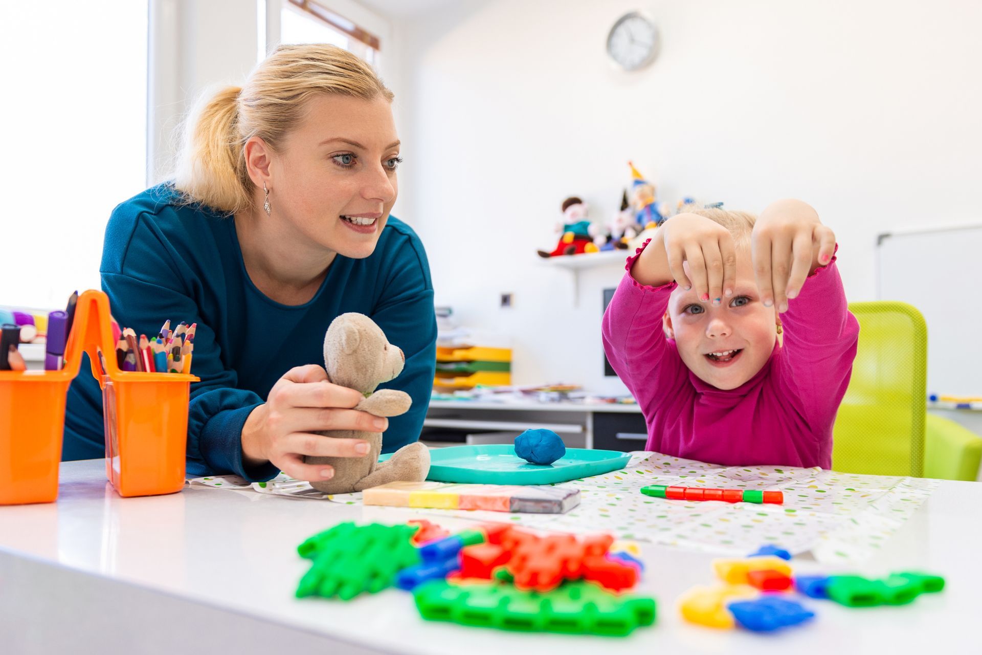 Woman and child at a table, playing with toys. The child is holding her hands up near her face.