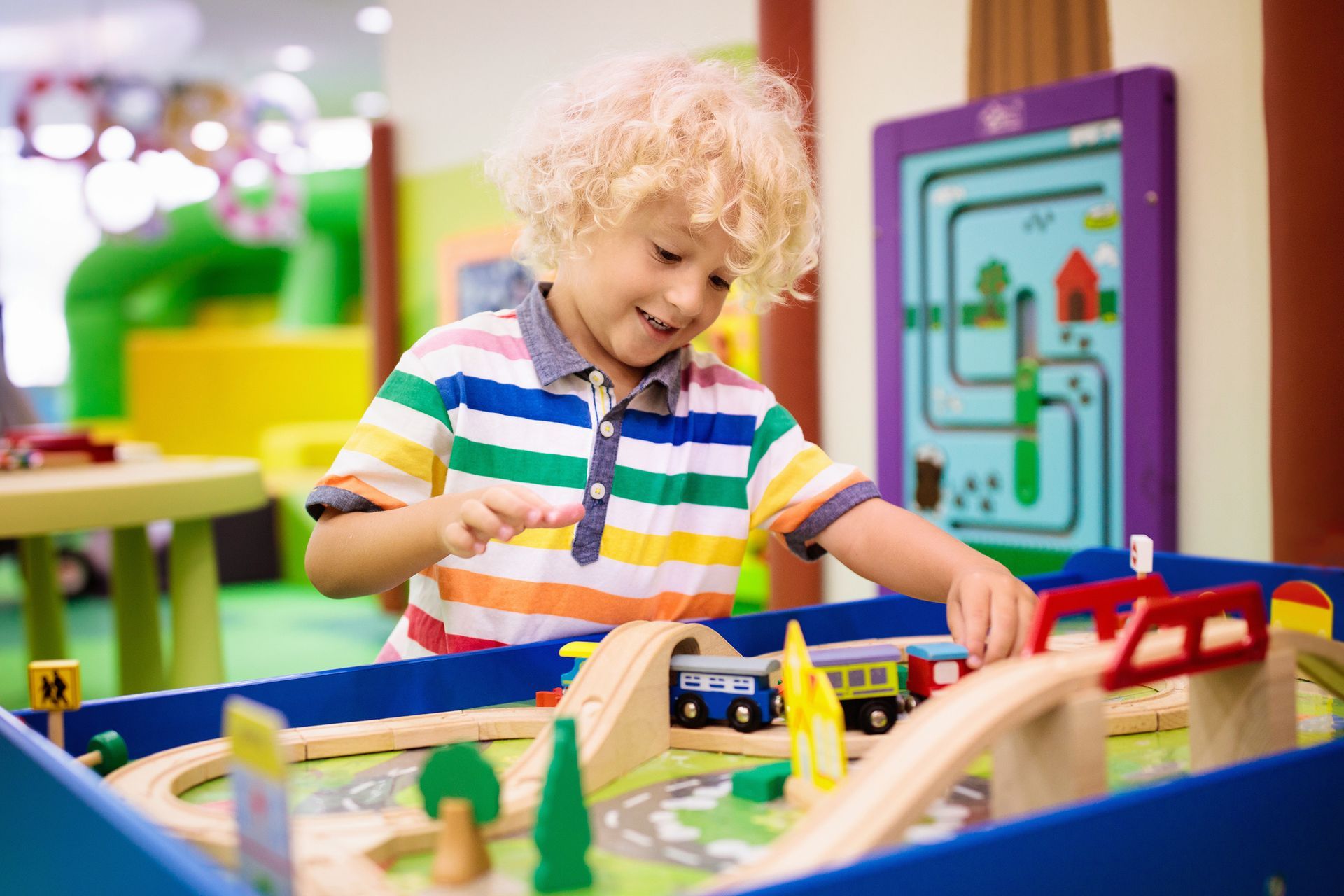 Boy smiles while playing with a wooden toy train set. Colorful shirt, indoor setting.
