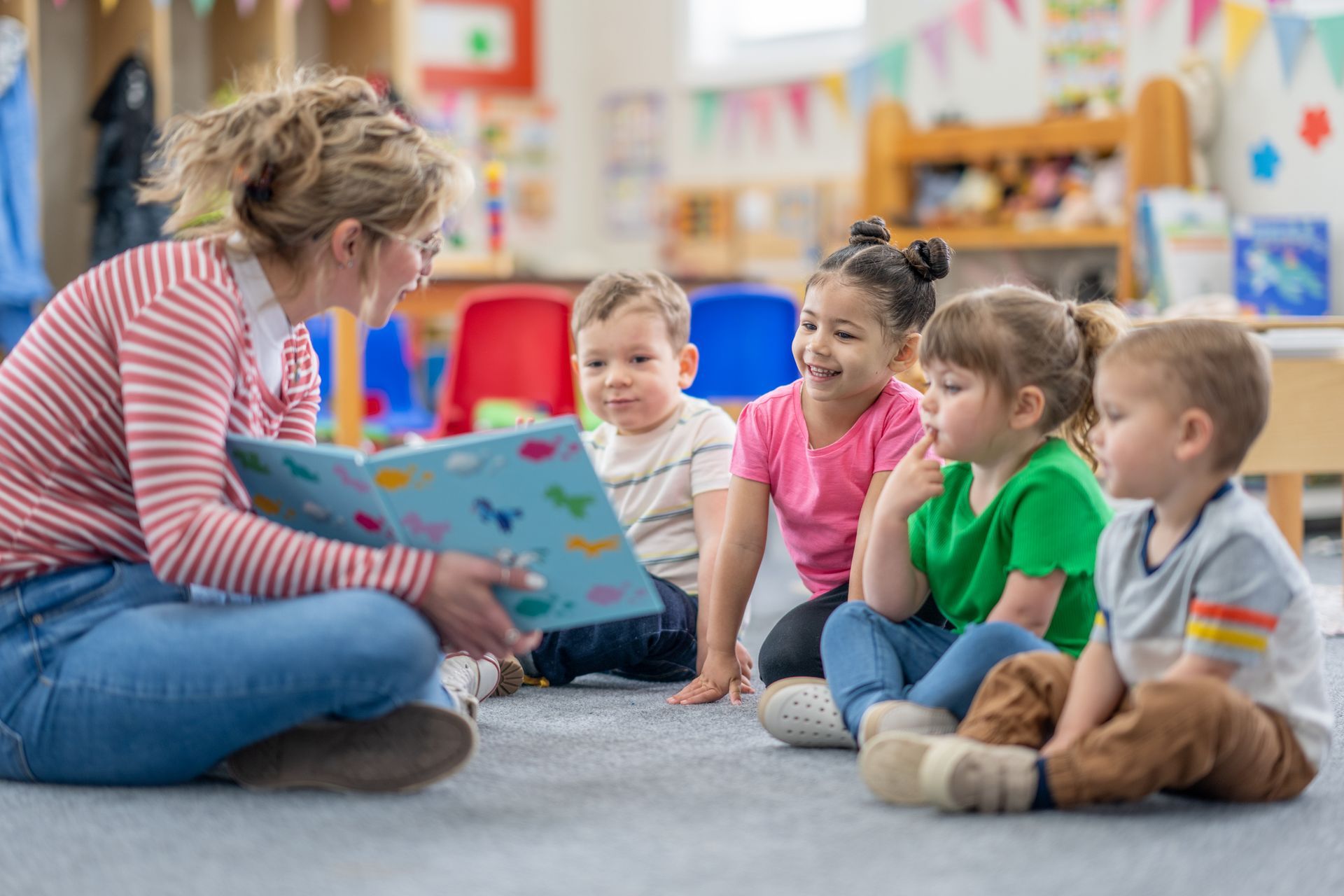 A teacher sits on the floor of her classroom with a group of students as she reads them a book.