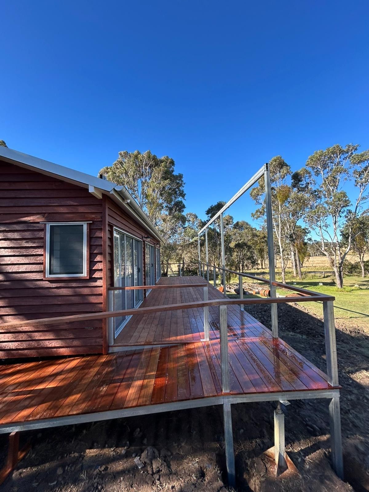 Wooden house deck with metal railing, red floorboards, and trees under a clear blue sky — Taylormade Constructions & Renovations In Kingaroy, QLD