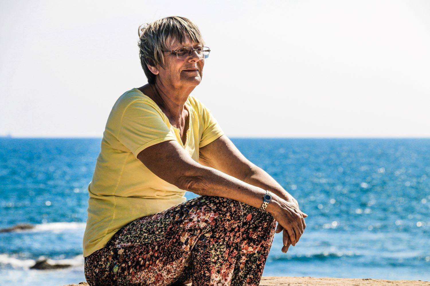 An elderly woman is sitting on the beach looking at the ocean.