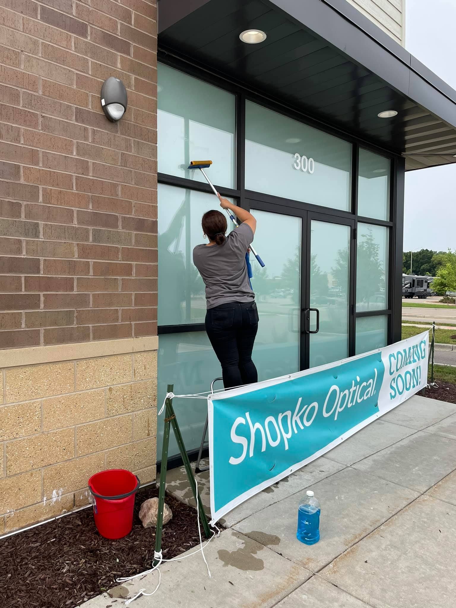 Person on a ladder washing windows of Shopko Optical storefront. Banner, bucket, and spray bottle visible.