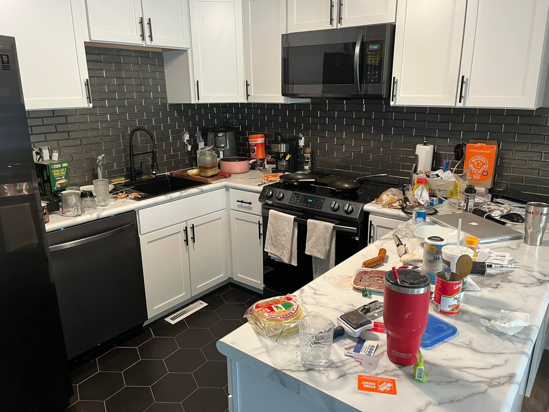 Cluttered kitchen with white cabinets, black backsplash, stainless appliances, and countertop island.
