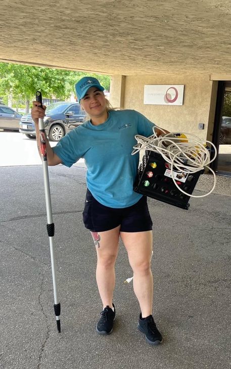 Woman holds walking stick and medical equipment outside a building, wearing blue shirt, shorts, and a hat.