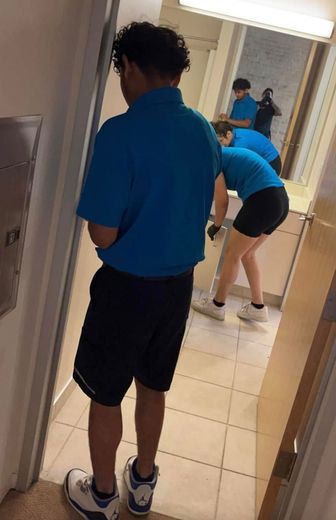 Man in blue shirt and shorts stands in bathroom doorway, watching people clean the sink.