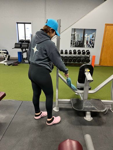 Woman in gym, adjusting exercise machine. She wears a blue cap, gray hoodie, black leggings, and pink Crocs.