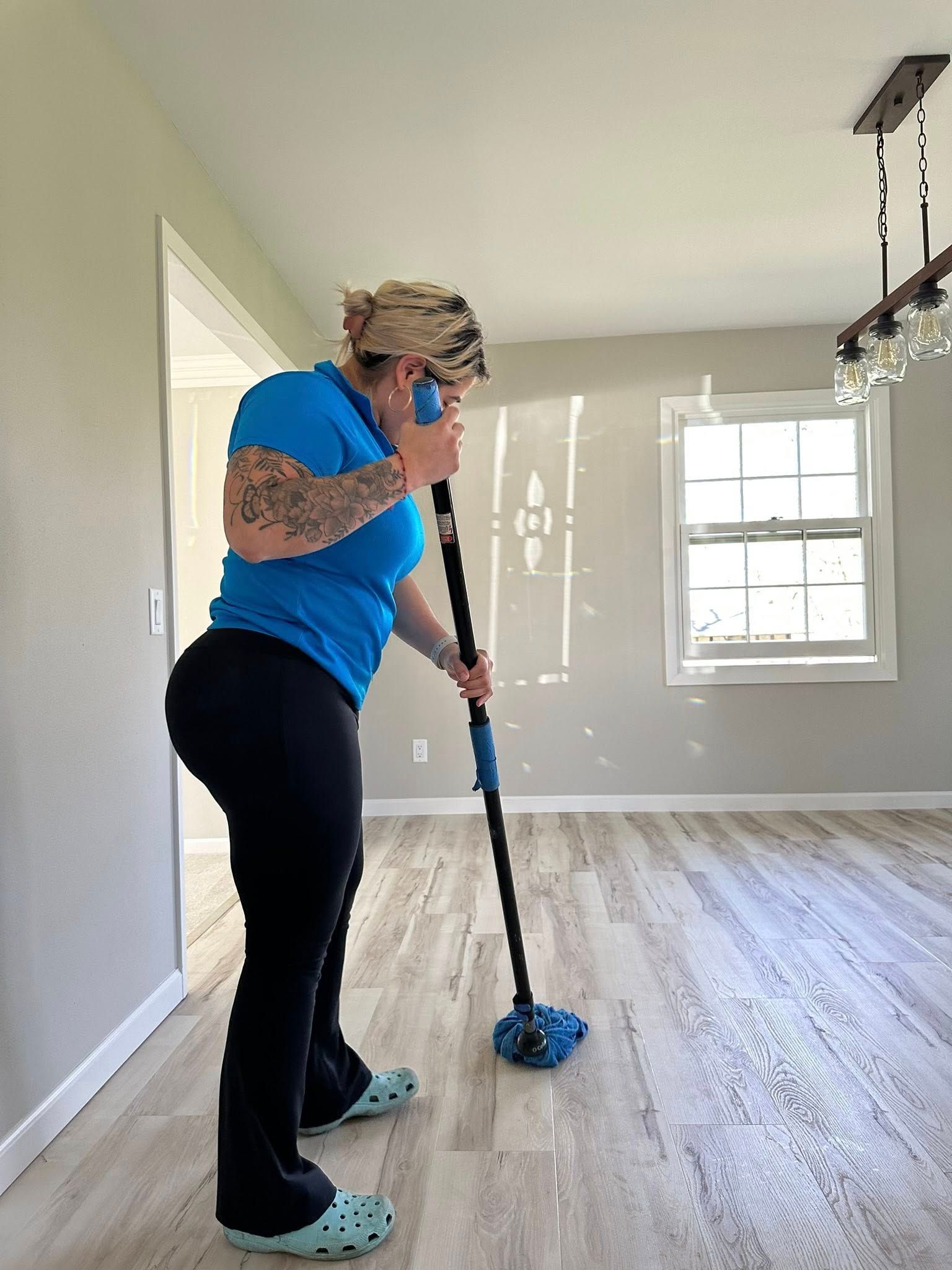 Person mopping a room while on the phone; wearing blue shirt, black pants, and blue clogs.